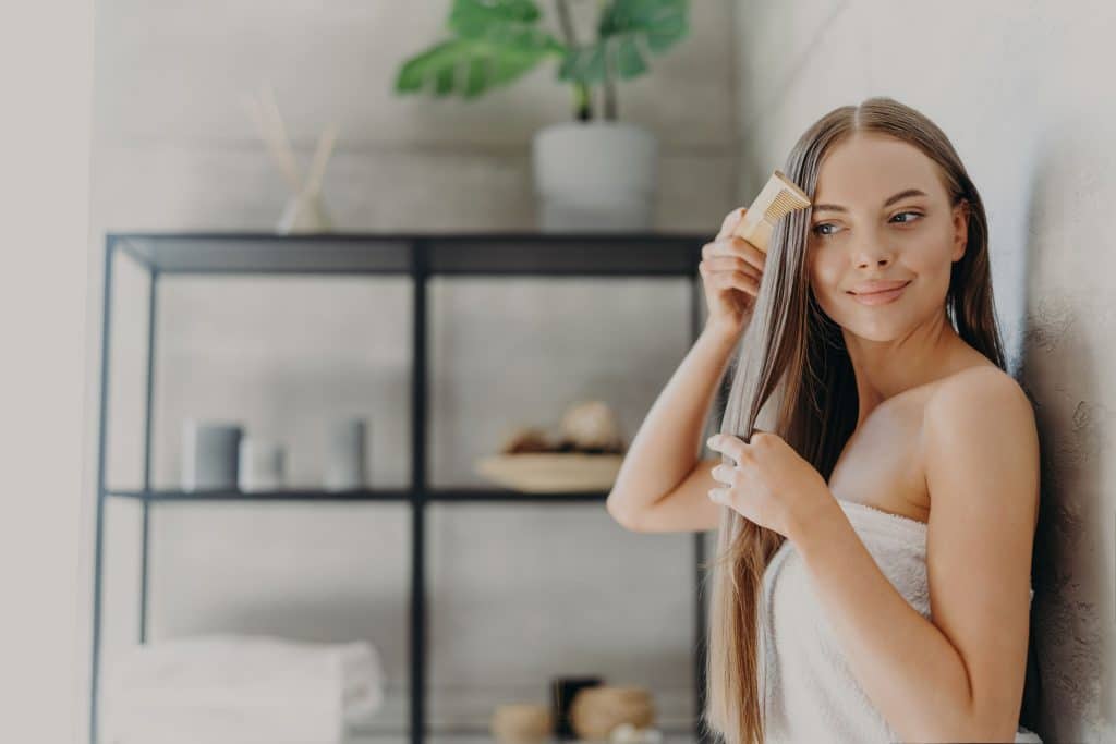 Woman in a bathroom combing her straight hair while wearing a towel
